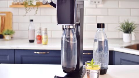 Ein Trinkwassersprudler steht in der Küche. Daneben steht eine weitere Flasche mit bereits aufgesprudeltem Wasser sowie ein Trinkglas mit Wasser und einer Zitronenscheibe drin.