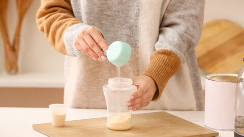 Eine Frau mischt mit einem Pulver eine Mahlzeit an. Es steht ein Gefäß auf einem Holzbrett und mit einem Löffel wird von der FrauPulver in das Gefäß gefüllt.