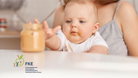 Kleines Baby sitzt auf dem Schoß der Mutter am Tisch und greift nach einen Gläschen mit Brei, in dem ein Löffel steckt.