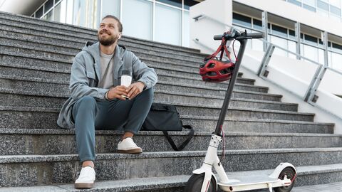Ein Mann sitzt mit einem Coffe-to-go-Becher vor einem Haus auf einer Treppe. Neben ihm steht ein E-Scooter. Am Lenker hängt ein Helm.