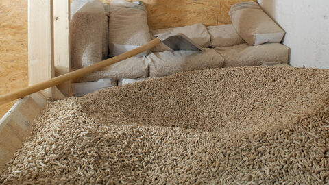 Ein Pelletlageraum mit losen Pellets im Vordergrund und im Hintergrund Pellets in durchsichtigen Kunstoffsäcken. Es liegt eine Schaufel auf den losen Pellets.